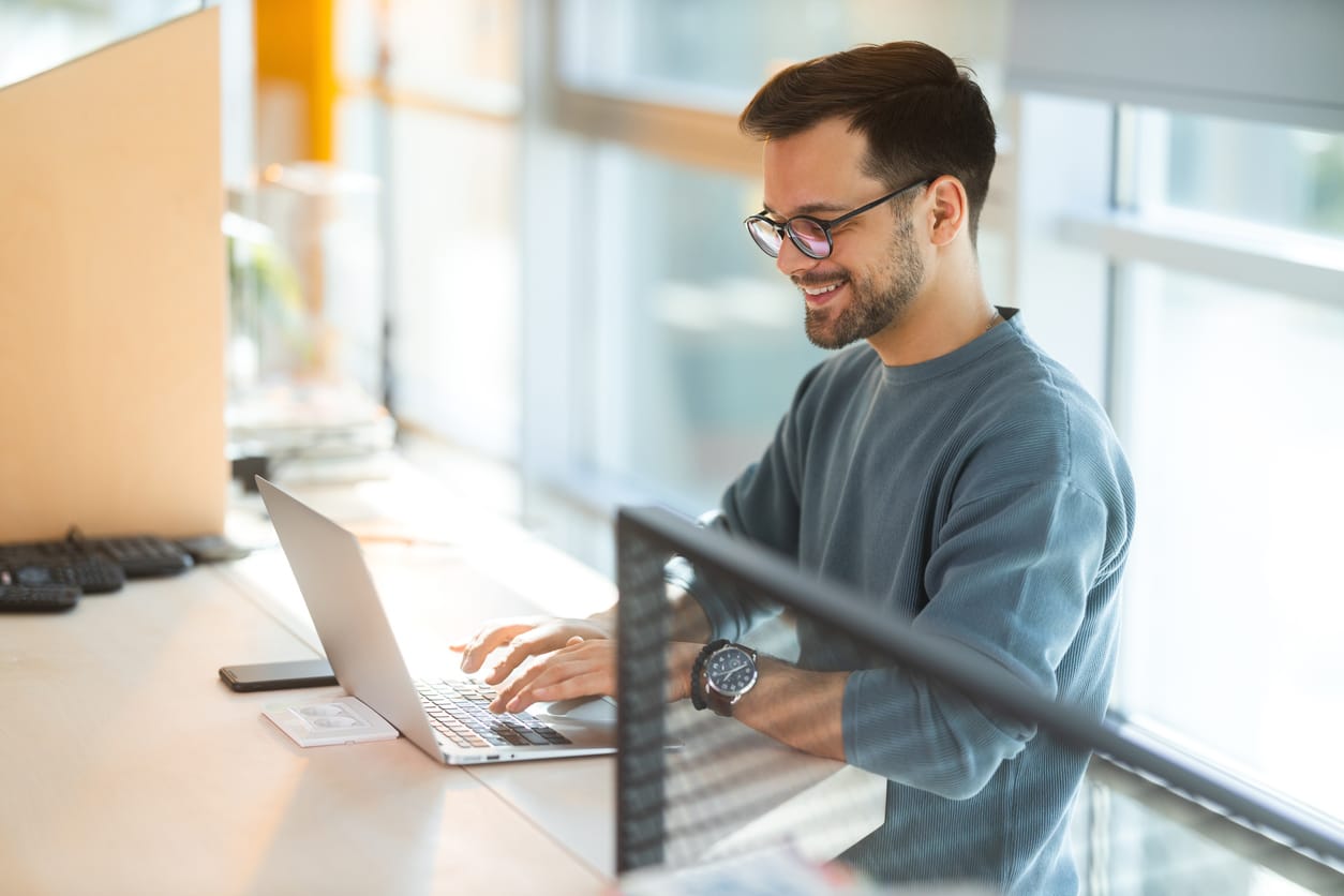 A smiling man working on a laptop in a bright office